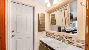 A bathroom with a white door, a large mirror with a decorative frame, a white sink, and a tiled backsplash. - Sunset Suite in Deer Harbor (Eastsound)