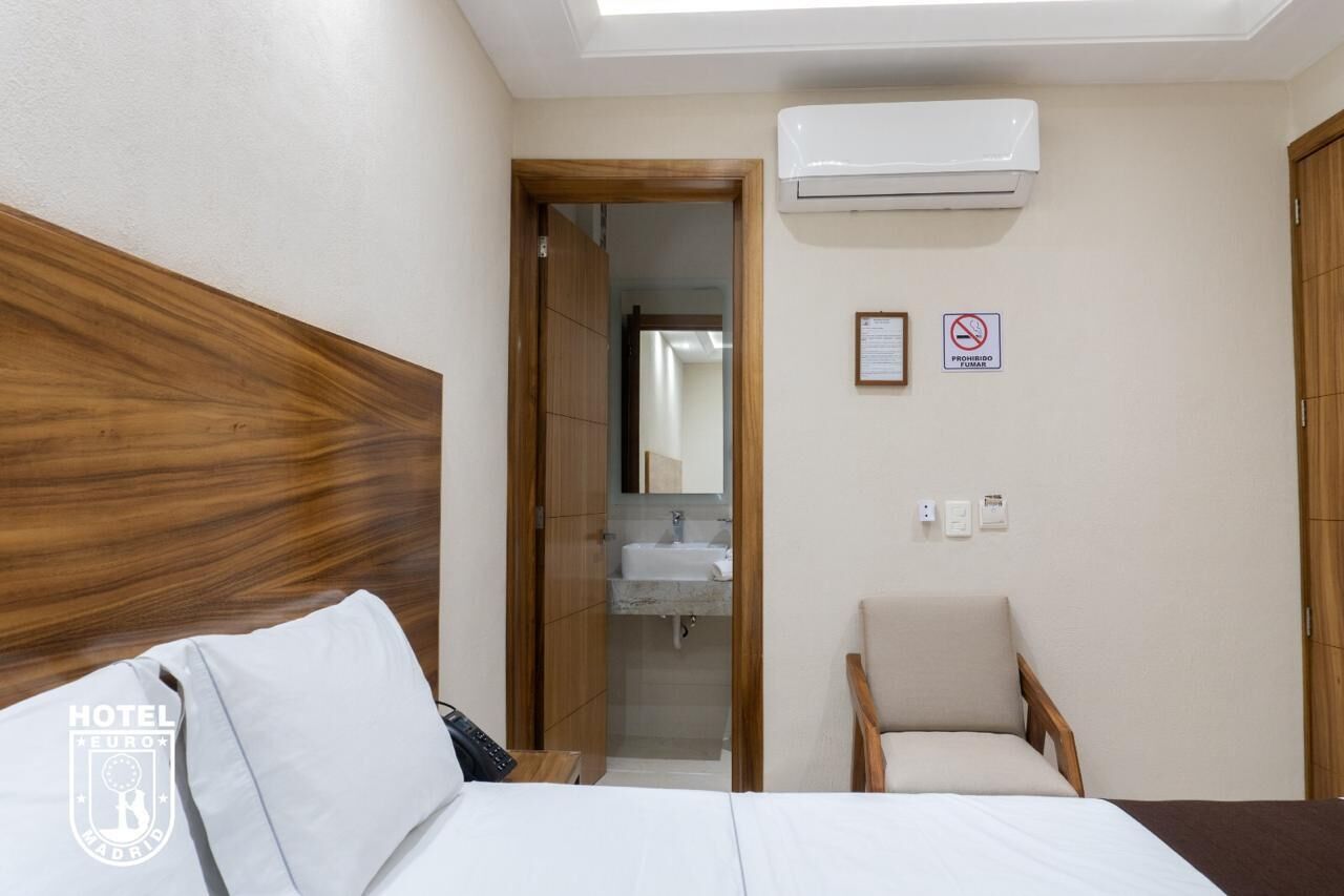 A hotel room with a wooden headboard, a bed with white bedding, a small armchair, a bathroom with a sink and mirror, and an air conditioning unit.
