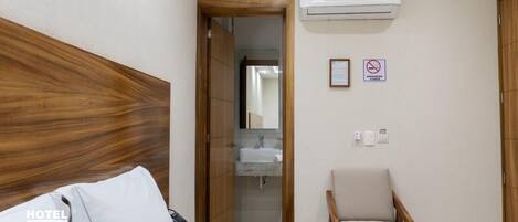A hotel room with a wooden headboard, a bed with white bedding, a small armchair, a bathroom with a sink and mirror, and an air conditioning unit.