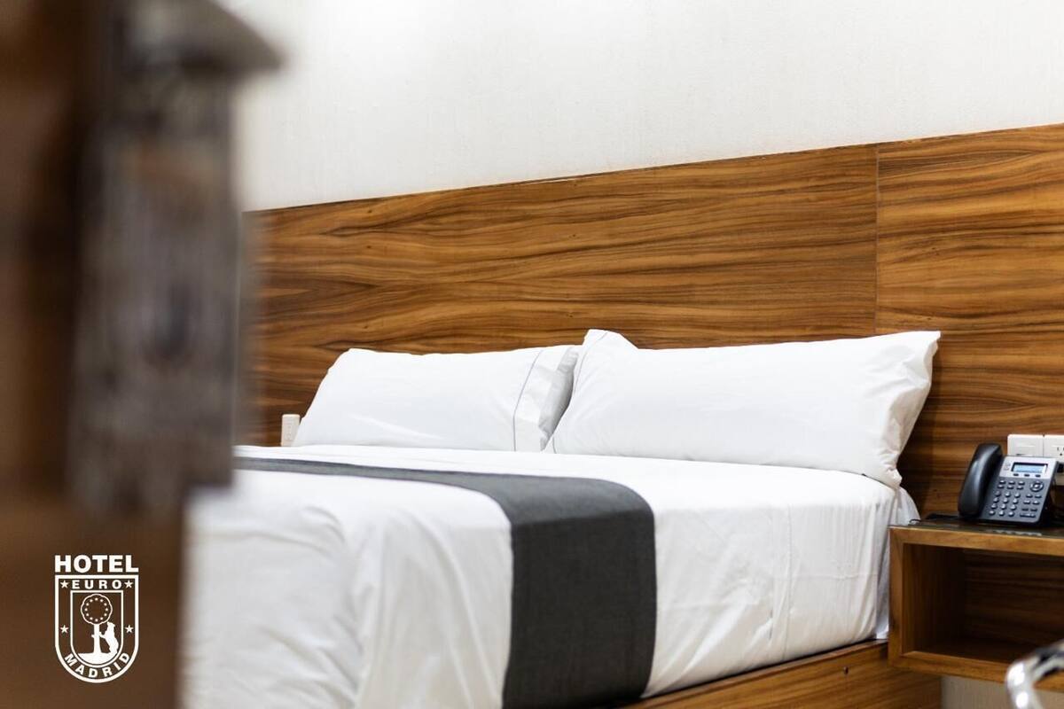 Ein Hotelzimmer mit einem hölzernen Kopfteil, weißer Bettwäsche, einem schwarzen Telefon auf dem Nachttisch und einem Logo an der Wand.