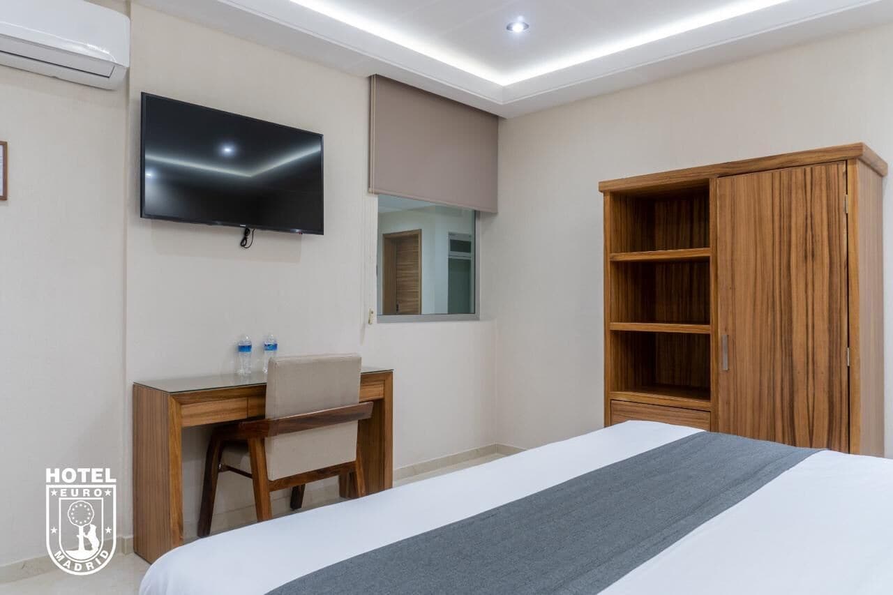 A hotel room with a bed, a desk with a chair, a television mounted on the wall, and a wardrobe.