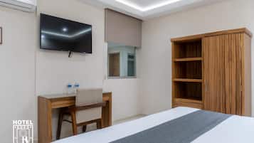 A hotel room with a bed, a desk with a chair, a television mounted on the wall, and a wardrobe.