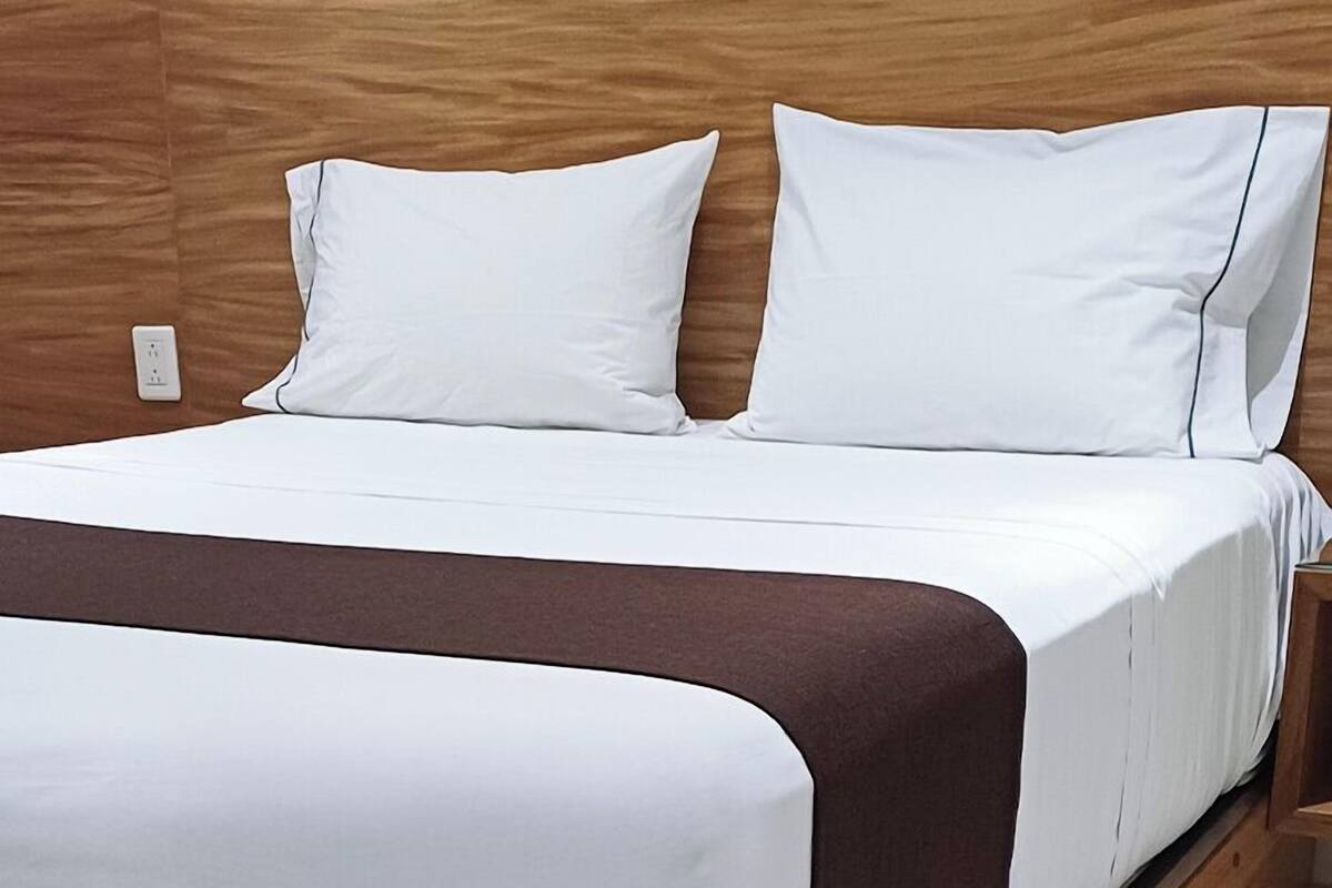 Ein ordentlich bezogenes Bett mit weißer Bettwäsche und braunen Akzenten, in einem Zimmer mit Holz-Kopfteil und weißer Wand.