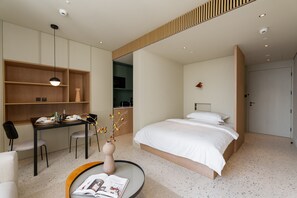 A modern hotel room with a large bed, a dining area with a table and chairs, a small kitchen, and a compact workspace.