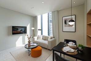 Un salon moderne avec un canapé, une table à manger pour deux personnes, un téléviseur fixé au mur et une grande fenêtre avec des rideaux.