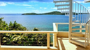 A spiral staircase leading to a balcony overlooking a clear blue sea with distant green hills.
