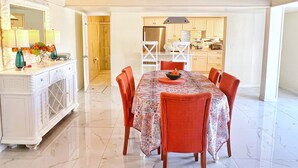 Een ruime eethoek met een houten plafond en een grote tafel, omringd door stoelen. In de achtergrond is een keuken zichtbaar.