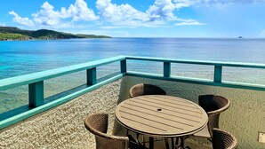 A seaside terrace with a table and chairs, overlooking a calm sea and distant hills.