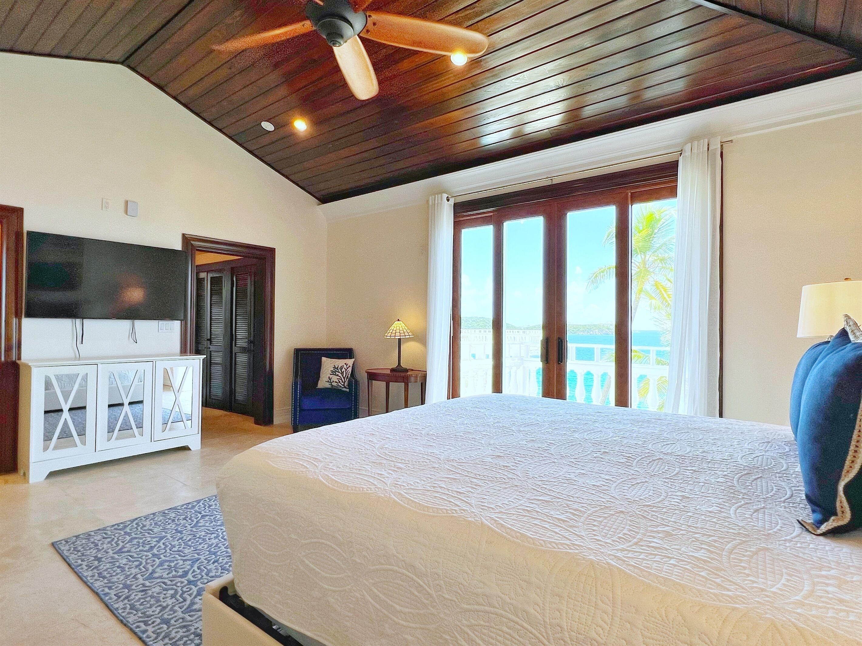 A spacious bedroom with a large bed, a ceiling fan, a TV, and a view of the outdoors through a sliding door.