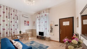 Una sala de estar con un sofá azul, una mesa de comedor, un televisor y un jarrón floral.
