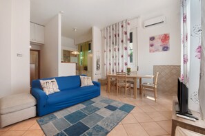 Un salon avec un canapé bleu, une table à manger avec des chaises, un téléviseur et, au fond, un petit coin cuisine.