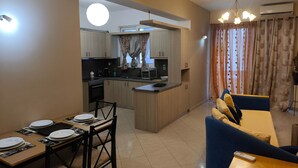 A modern kitchen with wooden cabinets, a dining area with a table set for four, and a living area with a sofa and a remote control on the table. - Odysseas Nea Chora Apartment with Sea View, Wi-Fi and Air Conditioning (Chania)