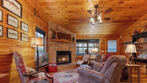 A cozy log cabin interior with a fireplace, wooden walls, a ceiling fan, and a comfortable seating area. - Mountain Views, Hot Tub + More! - Deer Crossing (Ellijay)