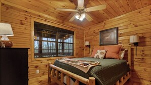 A wooden cabin bedroom with a bed, a window, a ceiling fan, and a framed picture on the wall. - Mountain Views, Hot Tub + More! - Deer Crossing (Ellijay)