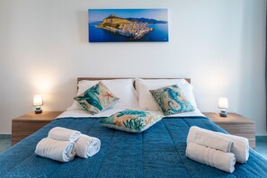 Ein Hotelzimmer mit Bett, Kissen, Nachttisch, Lampe und einem Gemälde einer Küstenstadt.