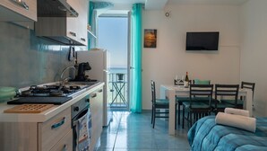 Una cocina funcional con estufa, lavabo y alacenas, un comedor con mesa y sillas, una cama con manta azul y un televisor en la pared.