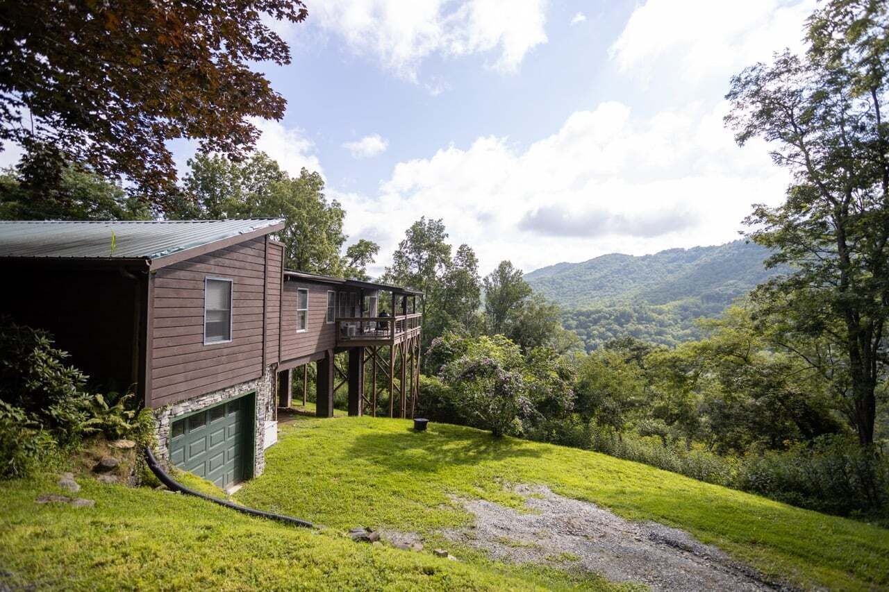 Una casa a due piani con un balcone, circondata da un prato e alberi, con vista su un paesaggio collinare.