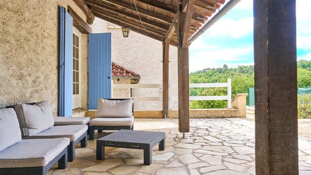 A covered outdoor seating area with stone flooring, a white fence, and a view of greenery.