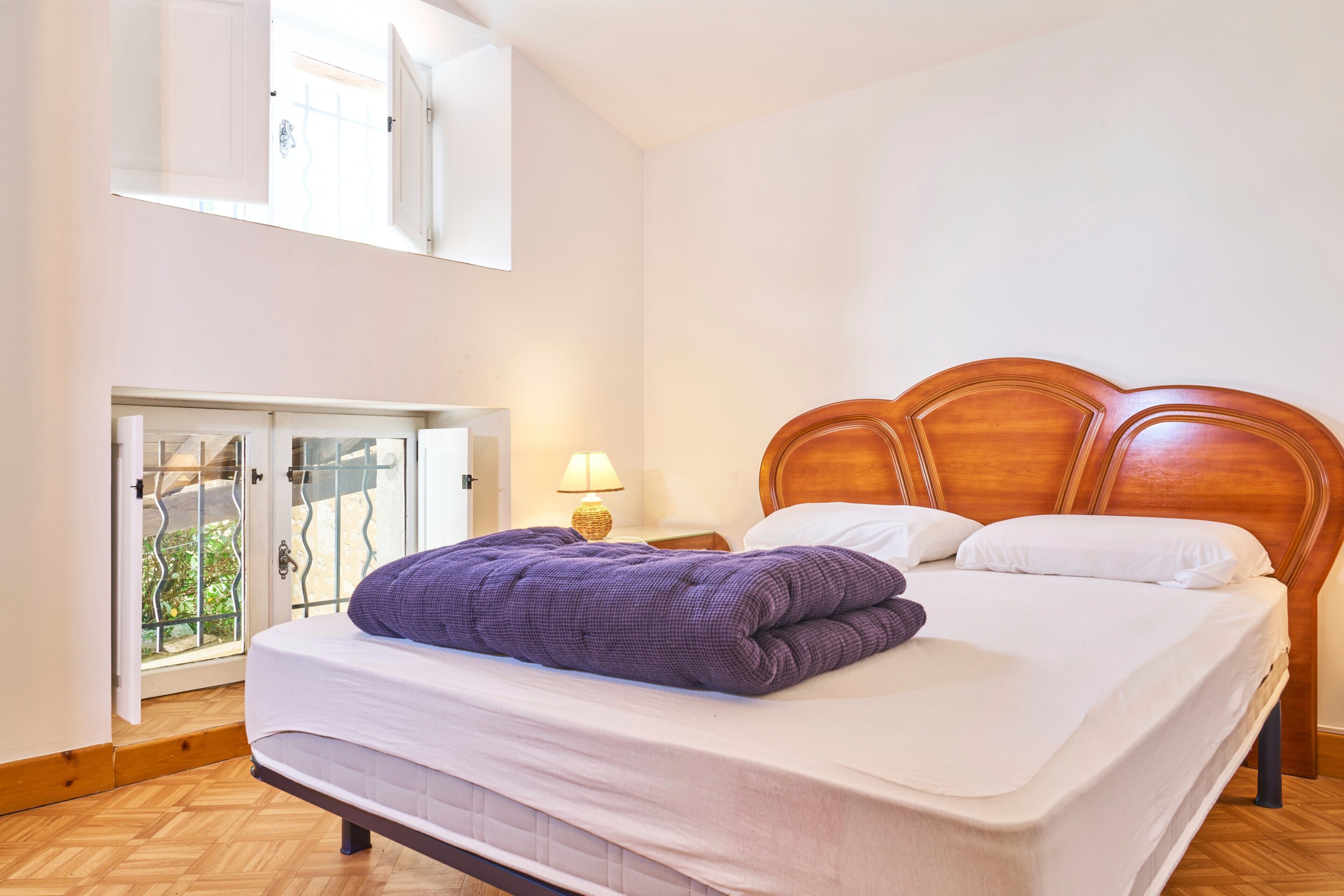 A bedroom with a wooden headboard, a bed with white bedding, a purple blanket, a nightstand with a lamp, and a sliding glass door leading to a balcony.