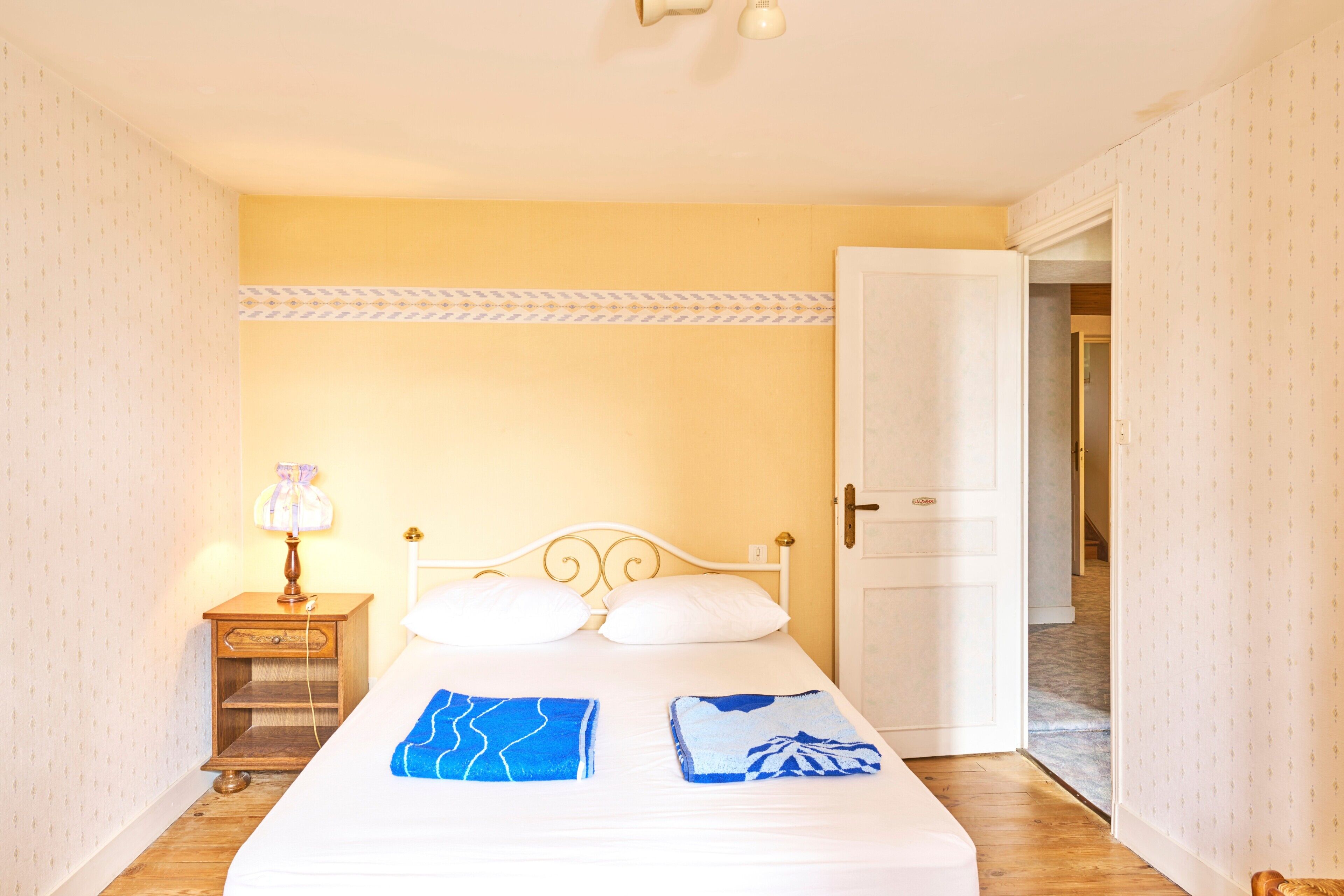 A bedroom with a bed, a nightstand with a lamp, and a door leading to another room.