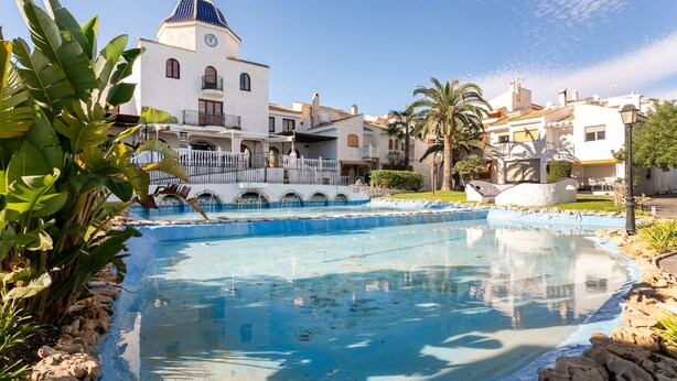 Una piscina con una fuente, rodeada de edificios blancos con balcones y una torre del reloj.