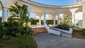 Un espacio al aire libre cubierto con columnas blancas, un sendero de ladrillo y abundante vegetación.