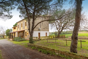 A two-story stone building with a yellow facade, a paved driveway, and a fenced garden area.