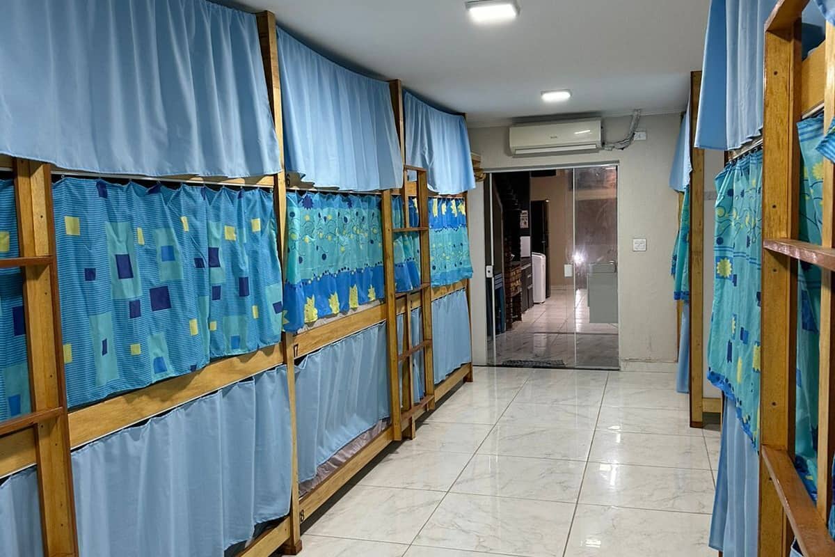 Un pasillo con literas cubiertas de cortinas azules, que lleva a una habitación con puerta de vidrio.