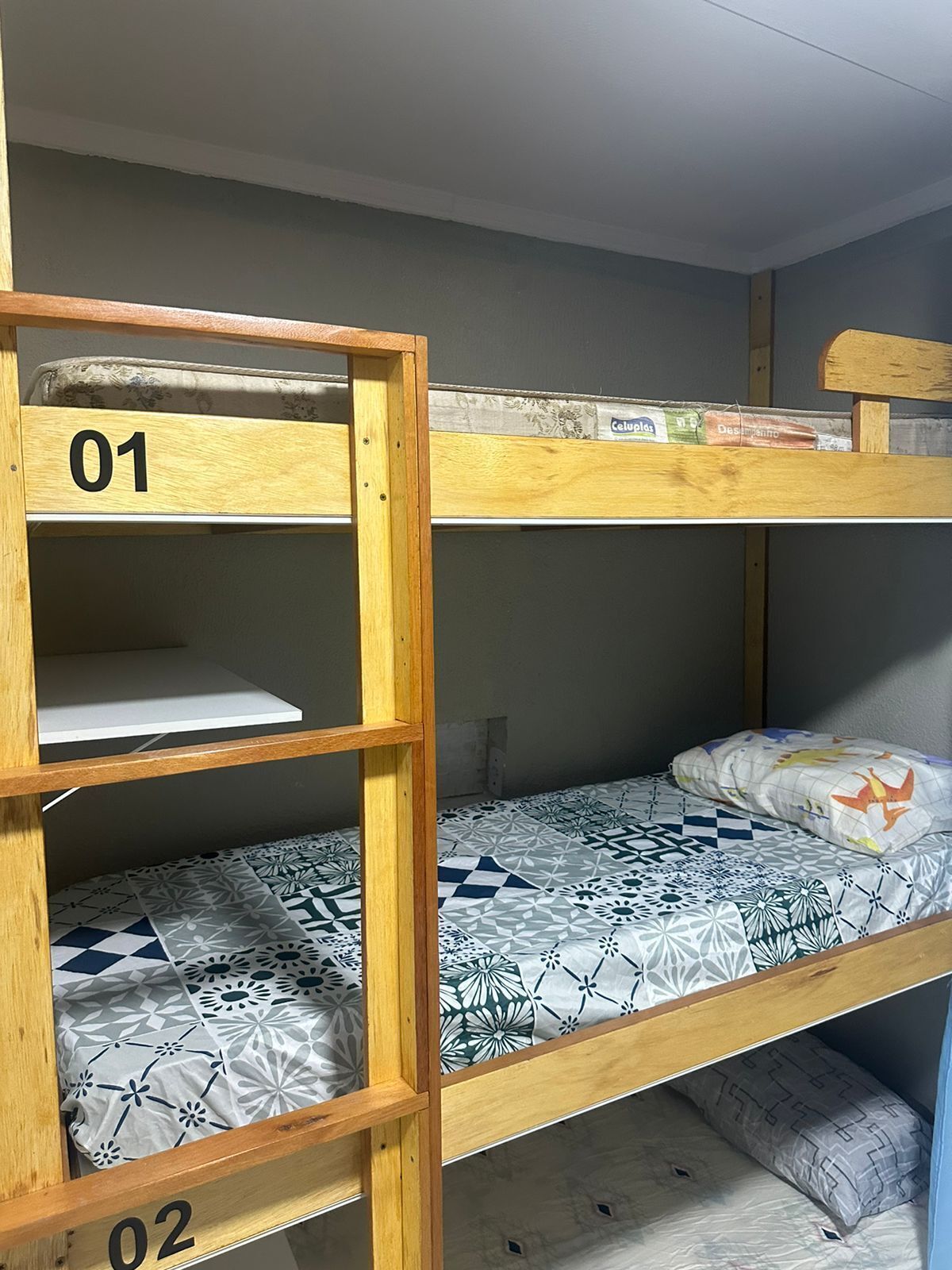 Letto a castello con due posti, ciascuno con struttura in legno e materasso. Il letto inferiore ha una coperta fantasia, mentre quello superiore presenta un materasso semplice.