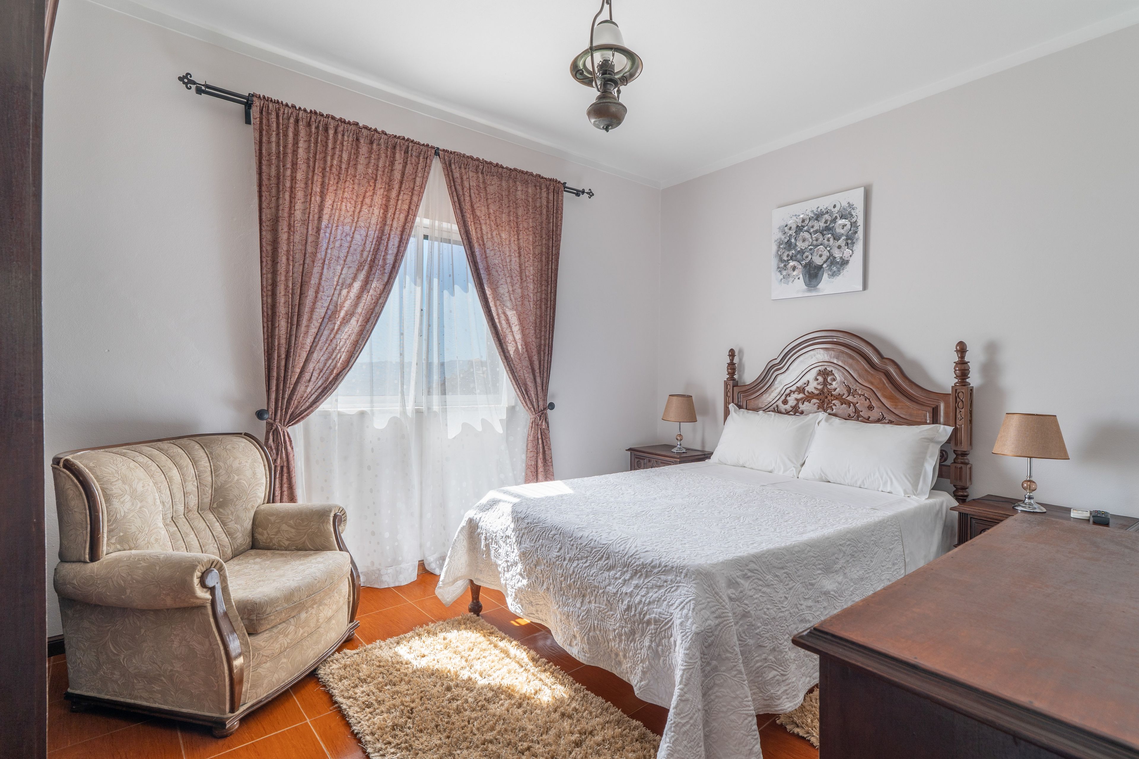 Un dormitorio con cama, una silla, una mesita de noche, una lámpara y un cuadro en la pared.