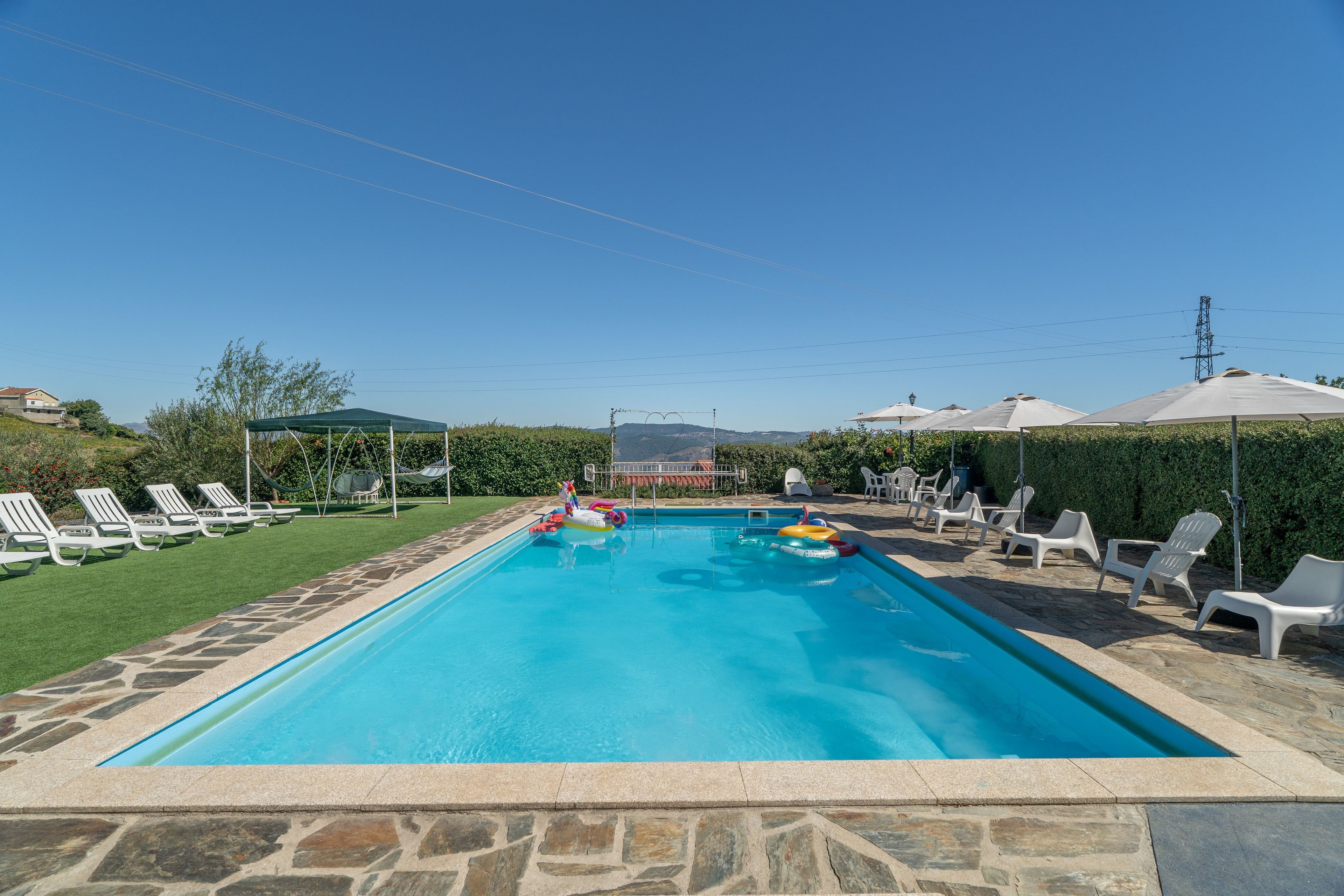 A rectangular swimming pool with a stone border, surrounded by white lounge chairs and umbrellas, with a clear sky above.