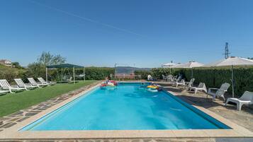 A rectangular swimming pool with a stone border, surrounded by white lounge chairs and umbrellas, with a clear sky above.