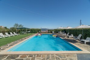 Una piscina rectangular con borde de piedra, rodeada de sillas de descanso y sombrillas, bajo un cielo despejado.