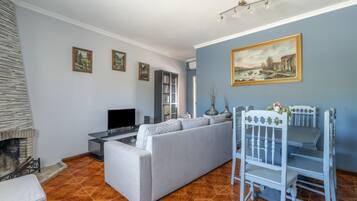 Un salon avec un canapé gris, un téléviseur à écran plat, une cheminée et un coin repas comprenant une table et des chaises blanches.