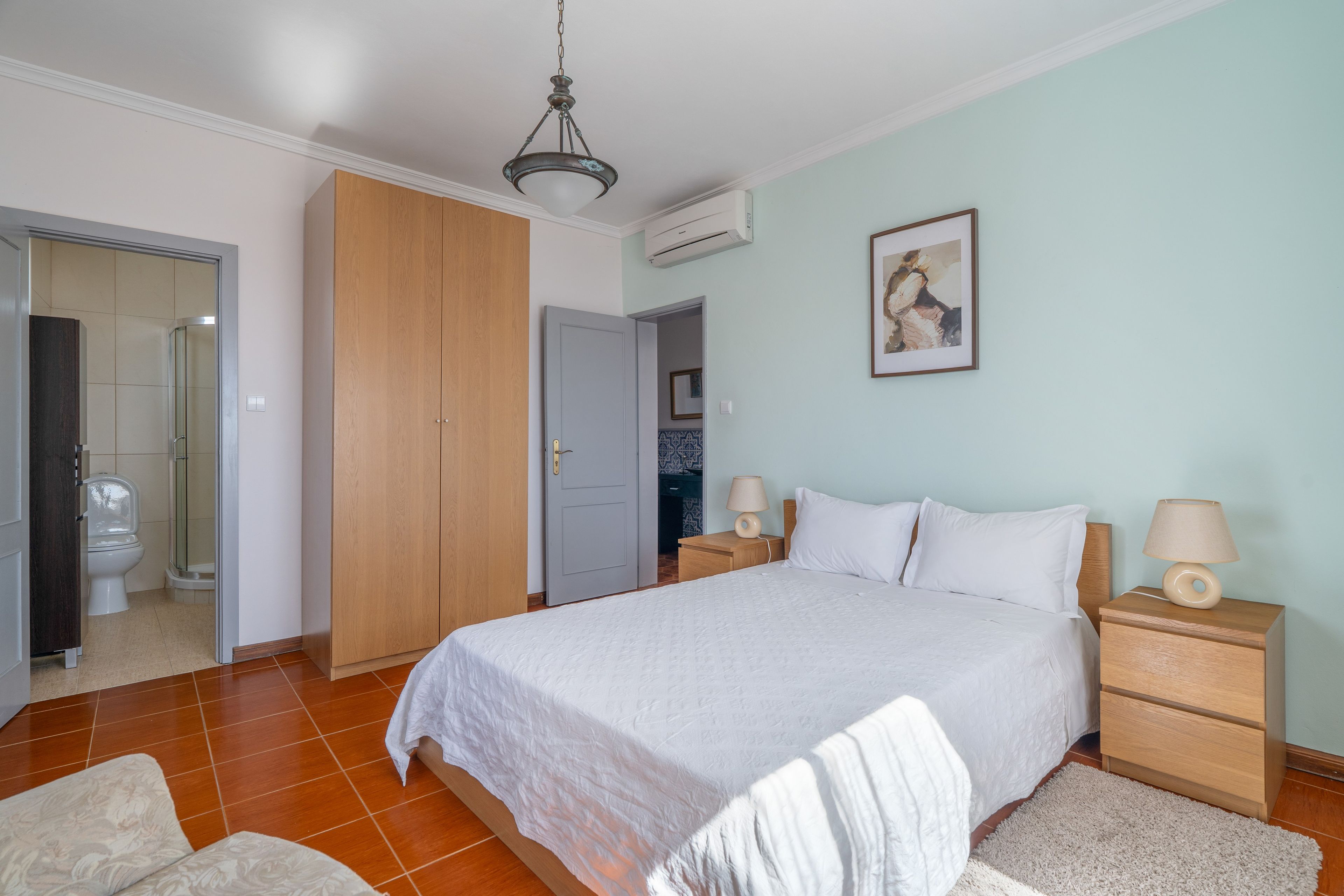 Un dormitorio con cama, mesitas de noche, lámpara de pared, un cuadro enmarcado y una puerta abierta que da a un baño.