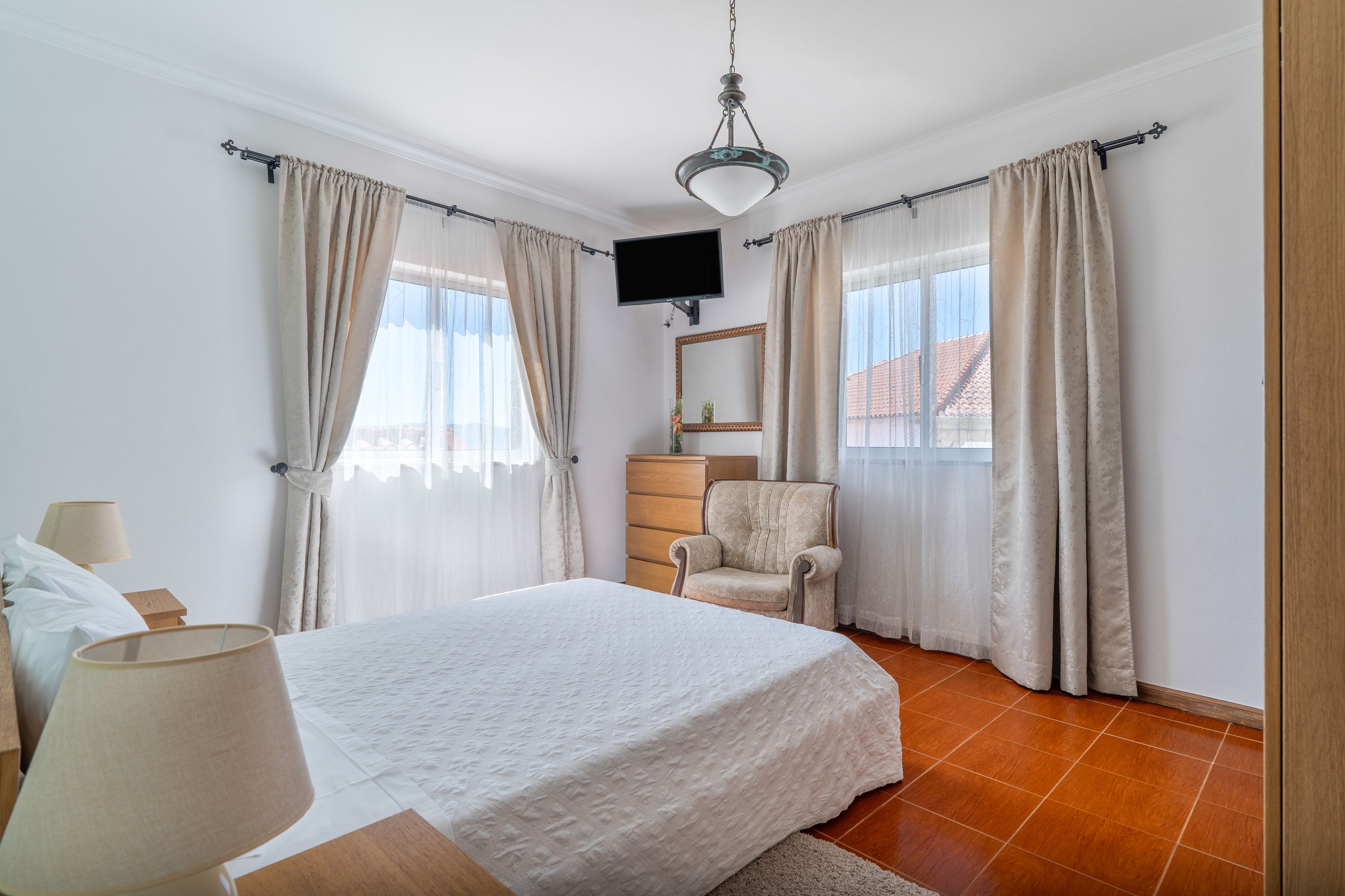 A bedroom with a bed, a chair, a TV mounted on the wall, and curtains on the windows.