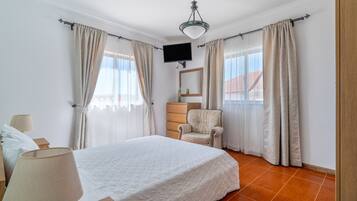 A bedroom with a bed, a chair, a TV mounted on the wall, and curtains on the windows.