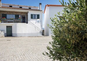 Un bâtiment à la façade blanche, doté d’un socle en pierre et d’un toit recouvert de tuiles. Un escalier mène à un balcon orné de fleurs.