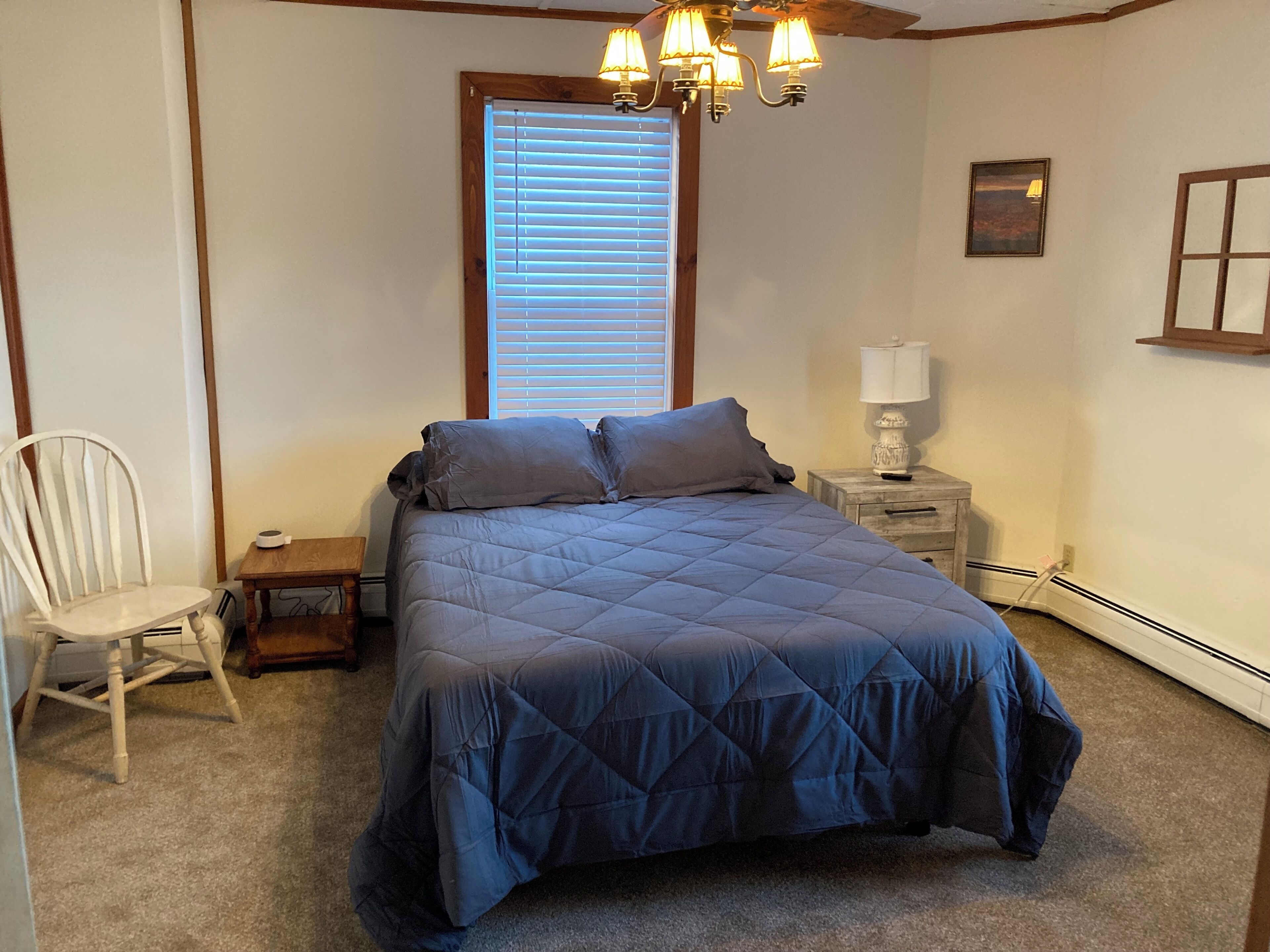 A bedroom with a bed, a chair, a nightstand, a lamp, a ceiling fan, and a window with blinds.