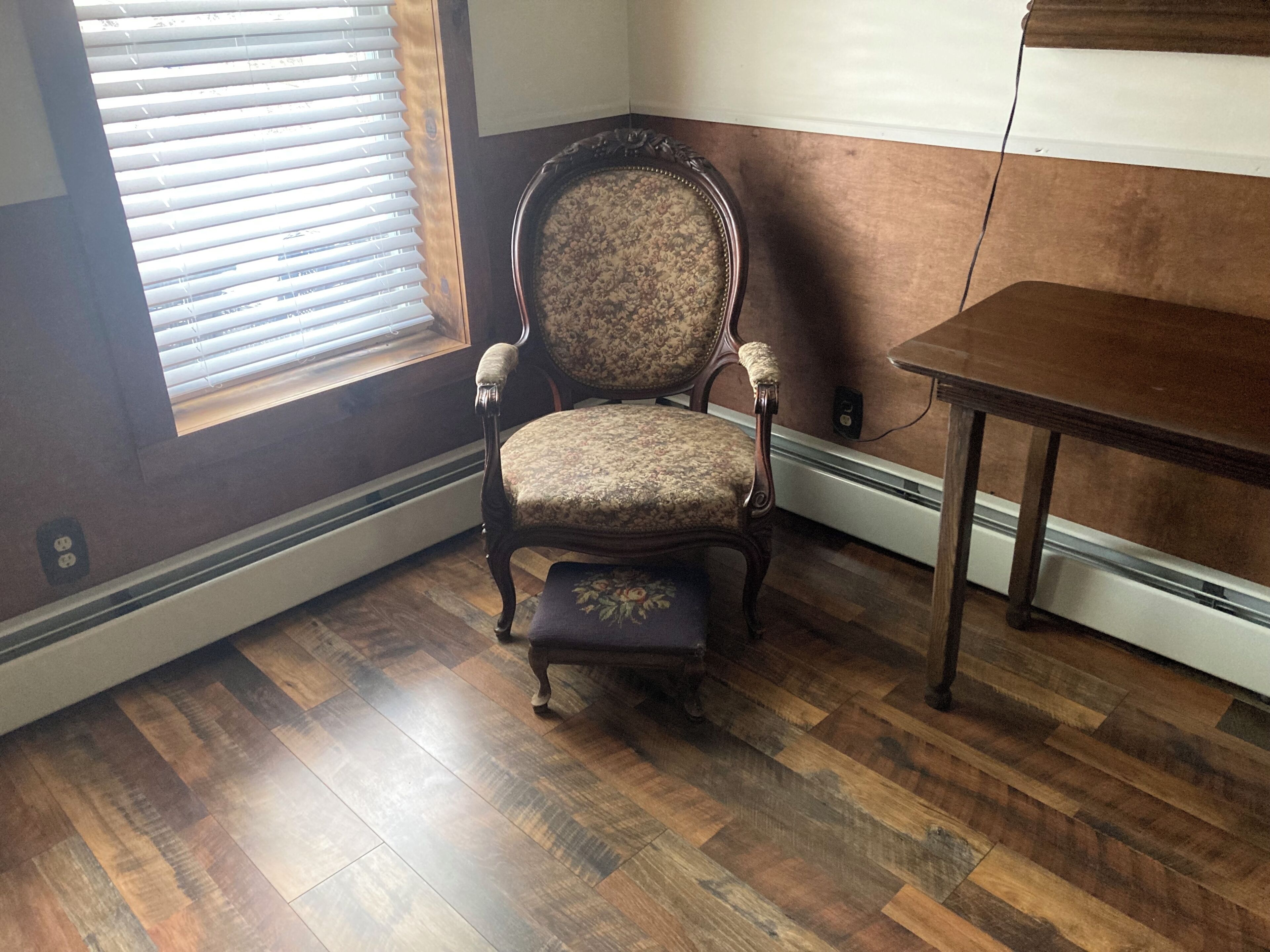 A room with a wooden floor, a chair with a floral pattern, a small table, and a window with blinds.