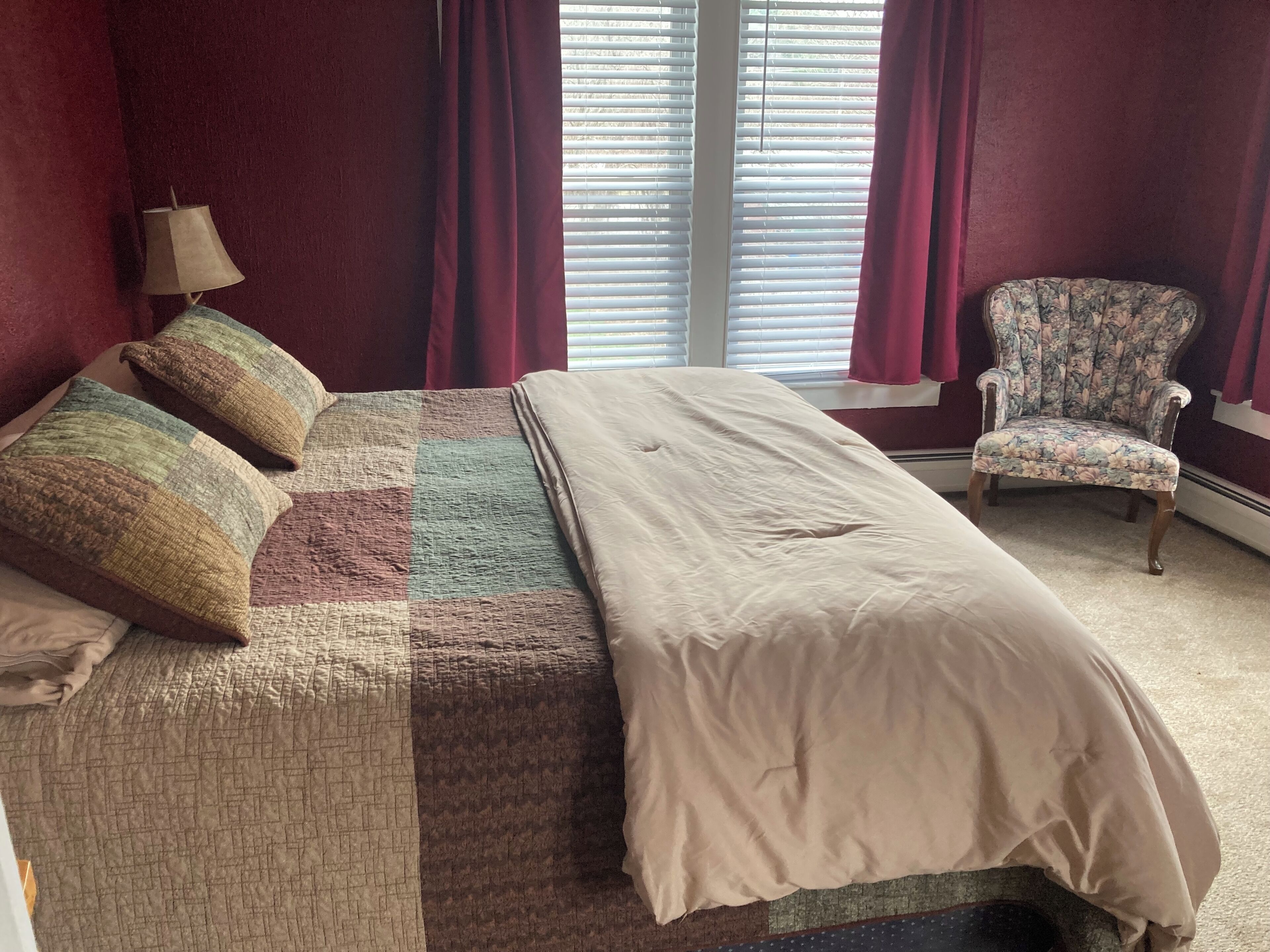 Una cama con colcha, almohadas, una lámpara, una silla y una ventana con persianas.
