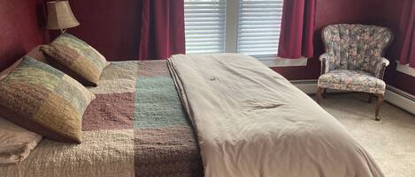 Una cama con colcha, almohadas, una lámpara, una silla y una ventana con persianas.