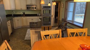 Una cucina con mobili bianchi, un piano di lavoro nero e un tavolo da pranzo in legno con quattro sedie. Si notano due forni, un forno a microonde e un frigorifero.