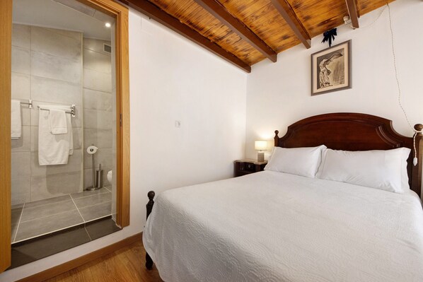 A bedroom with a wooden headboard, a bed with white bedding, a nightstand with a lamp, and a shower area with a glass door. - Romantic Room – mountain view, Wi-Fi and air conditioning (Provesende)