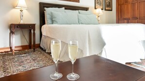 A neatly made bed with a white duvet and light blue pillows, two bedside lamps, a wooden nightstand with a lamp, and two glasses of white wine on a table.