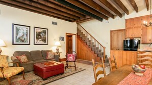 A living room with a sofa, ottoman, and chairs, a dining table with a rug, a staircase, wooden cabinets, and a TV.