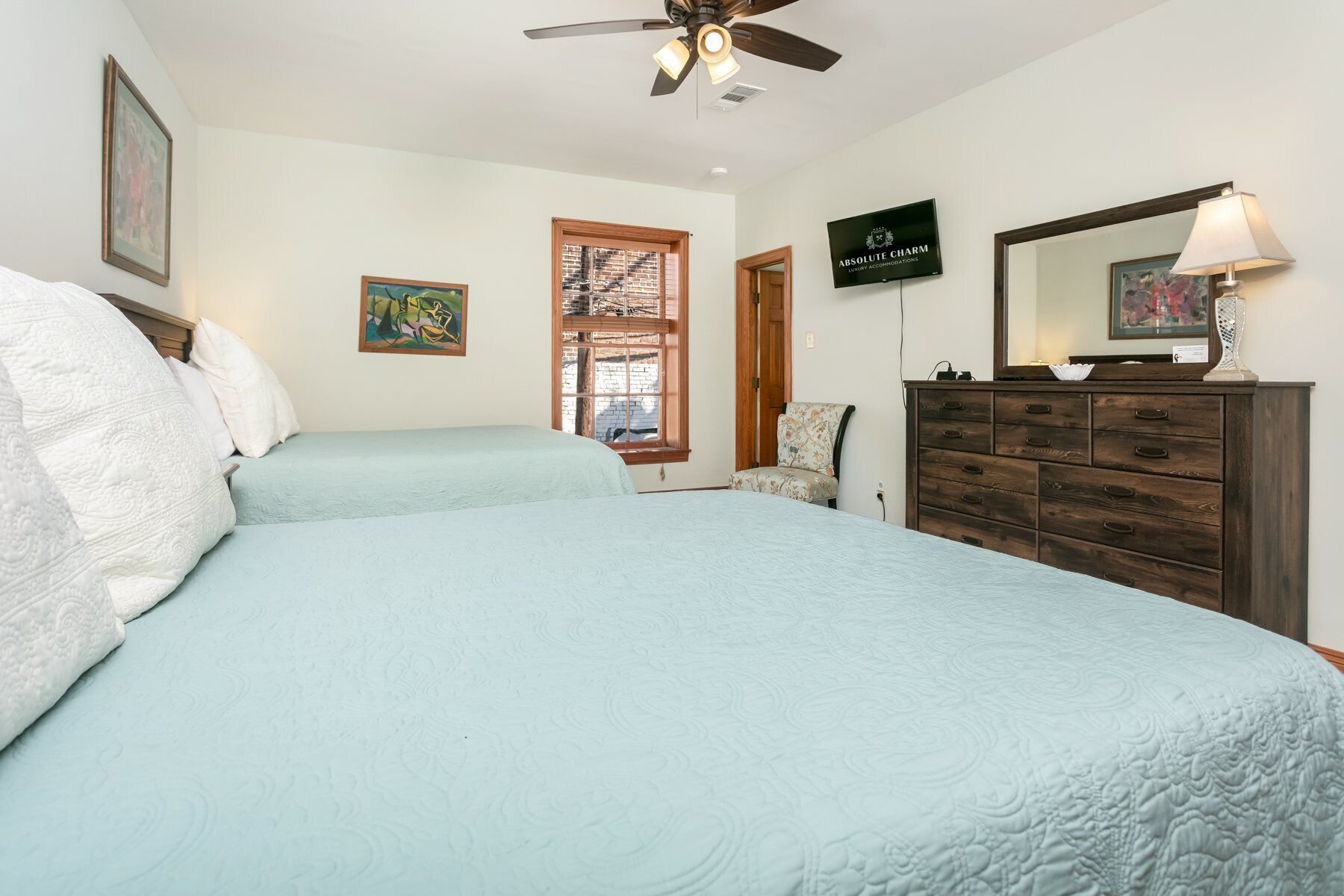 A bedroom with two beds, a ceiling fan, a wooden dresser, and a television displaying 'ABSOLUTE CHARM LUXURY ACCOMMODATIONS'.