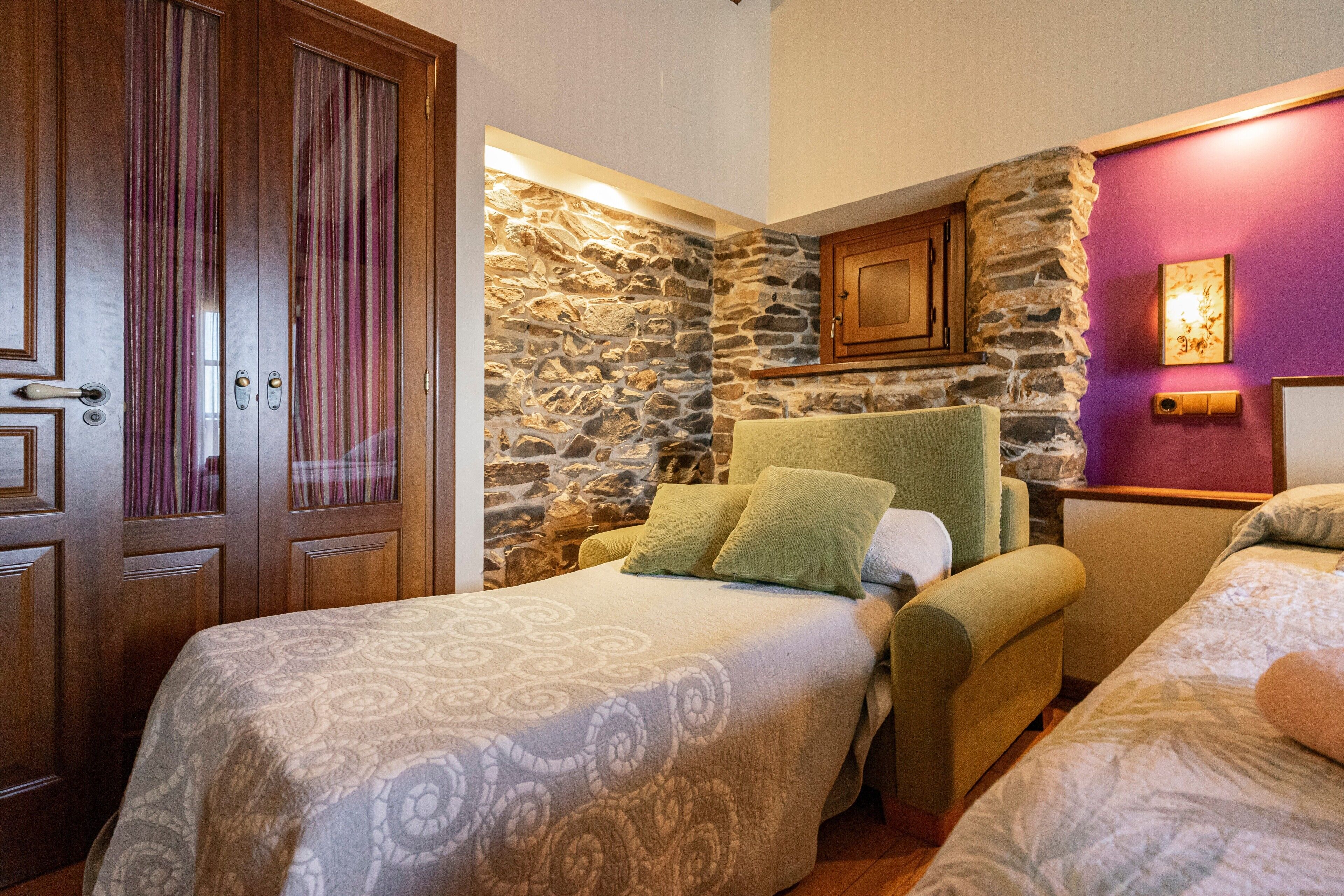 A room with two beds, a stone wall, wooden cabinets, and a green armchair.