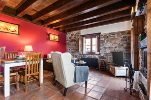 Una habitación acogedora con pared de piedra, vigas de madera, chimenea, televisión y comedor con sillas y mesa.