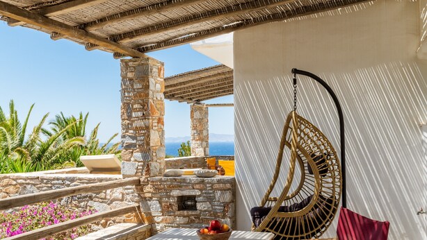 A stone balcony with a wicker chair, a table with a bowl of fruit, and a view of the sea.